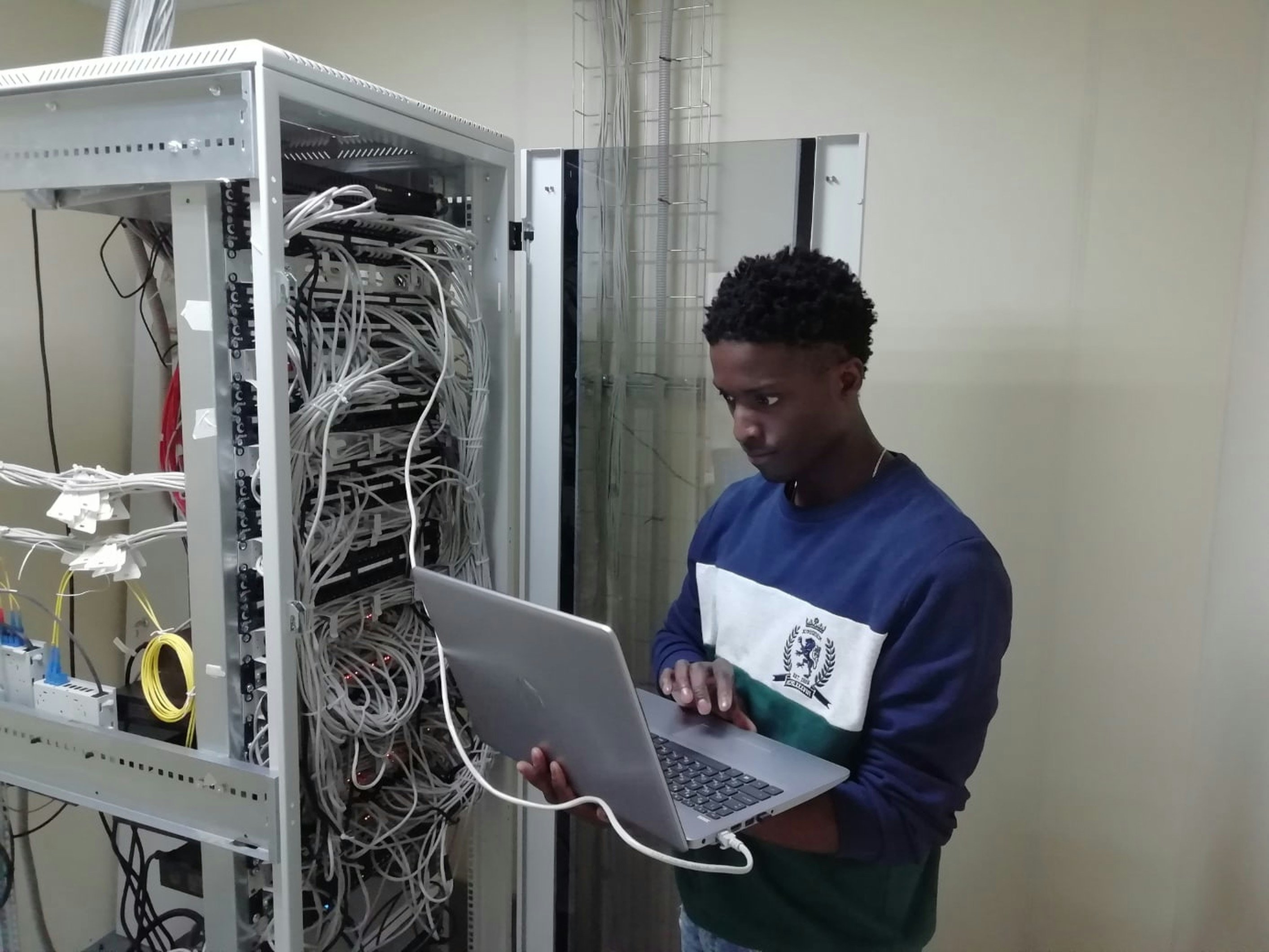 Person working on a laptop next to a server rack full of cables in a data center.