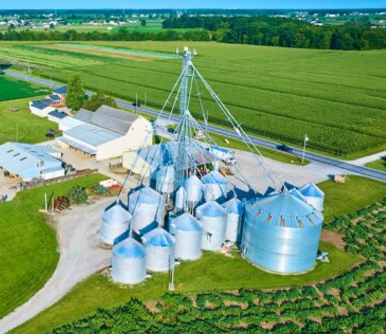 Aerial view of a large industrial farm complex with multiple silos, surrounded by lush green fields and a road.