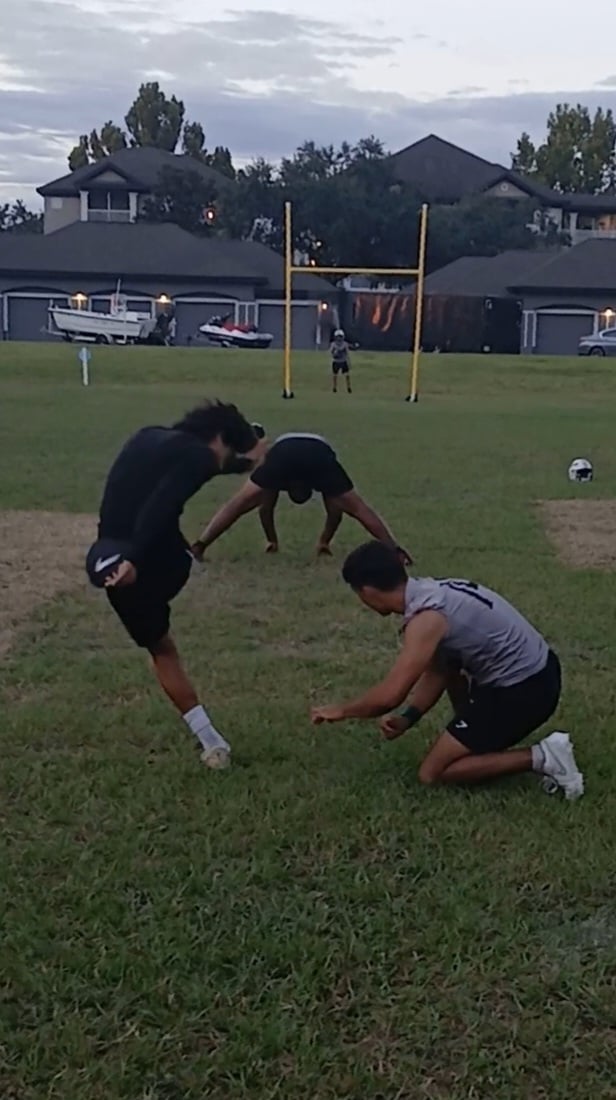 Three young men practicing football on a grassy field, focusing on kicking technique with a goalpost in the background.