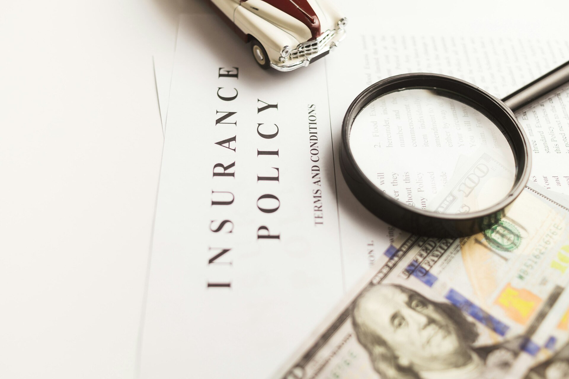 Close-up of an insurance policy document with a magnifying glass, toy car, and a hundred-dollar bill.