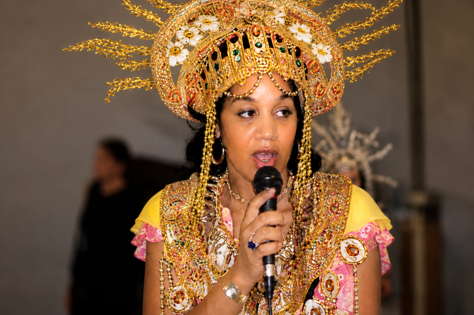 Three people engaging in a creative activity at a table, with one person having face paint and a colorful outfit, another person interacting, and a child in the background. Three people engaging in a creative activity at a table, with one person having face paint and a colorful outfit, another person interacting, and a child in the background.