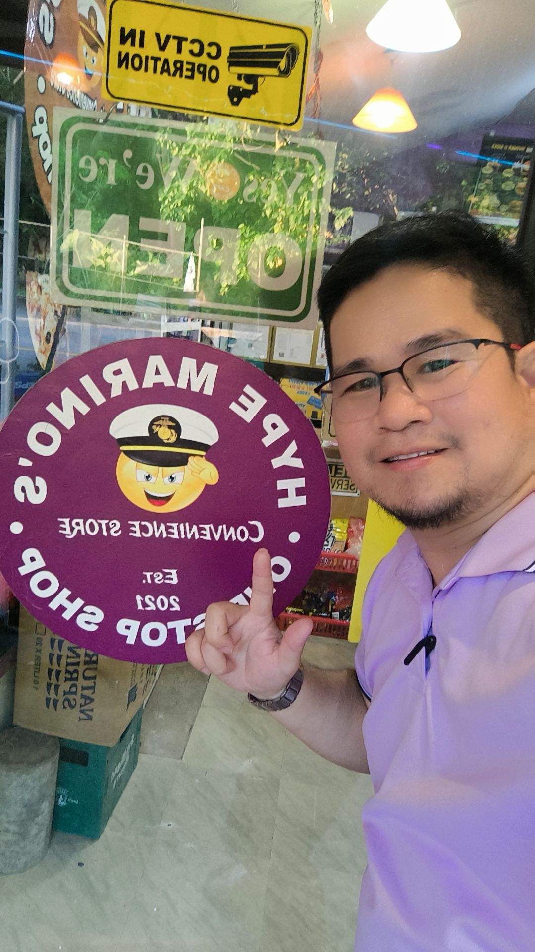 Man in glasses pointing at a convenience store sign with CCTV operation notice and open sign reflected on the glass door.