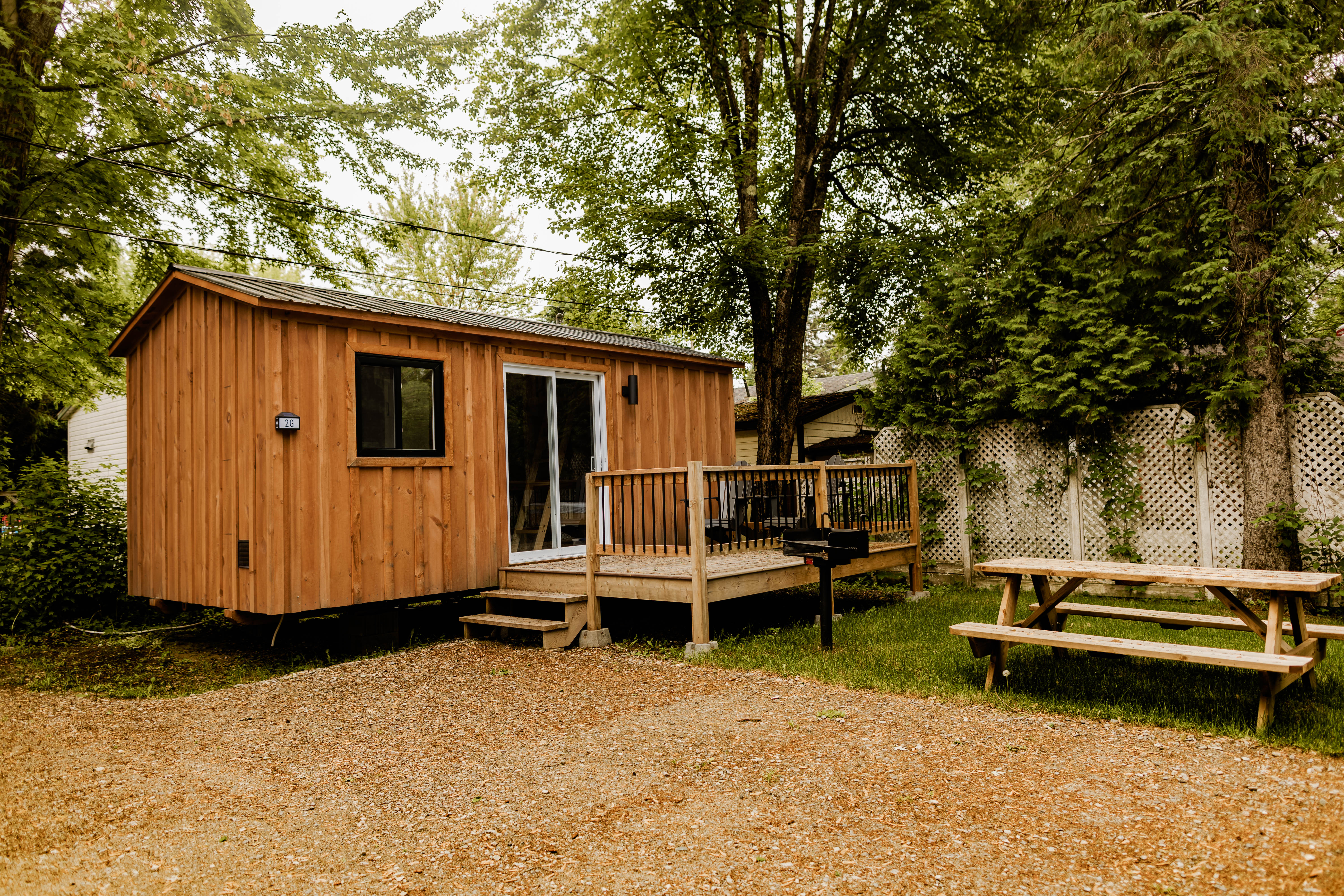 Camping en montagne avec tentes, cabane en bois, rivière, arbres verts et montagnes en arrière-plan par une journée ensoleillée.