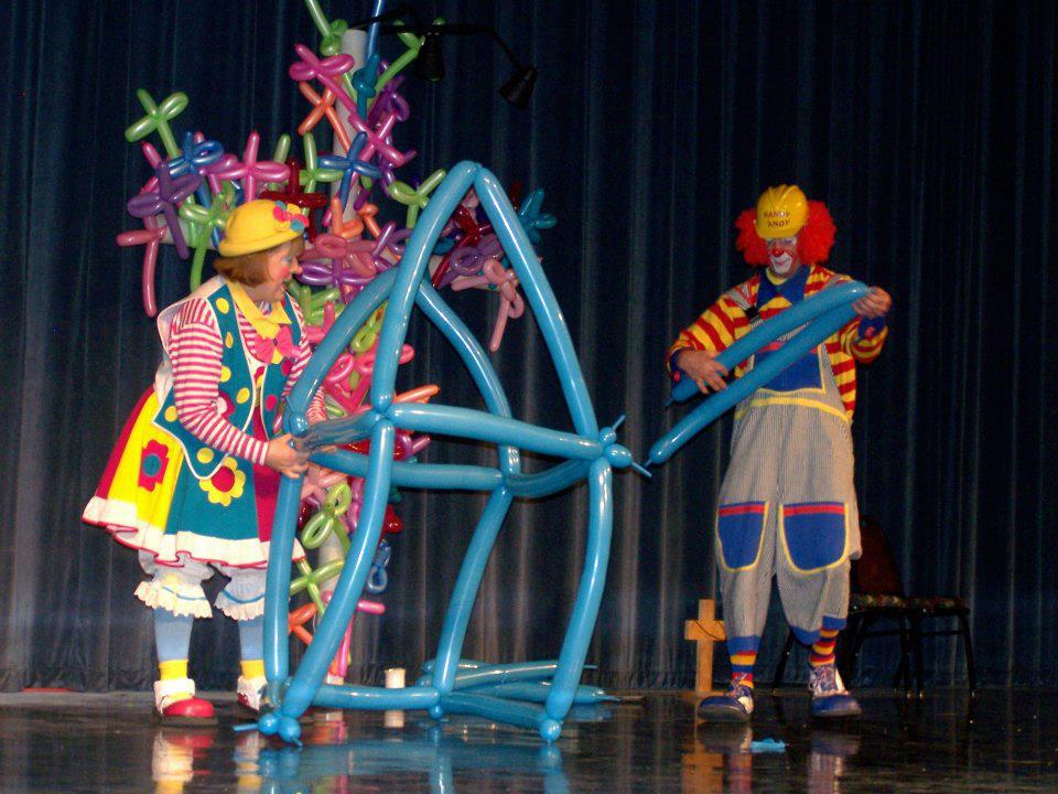 Cheerful clown with rainbow wig and face paint giving thumbs up surrounded by colorful balloons.