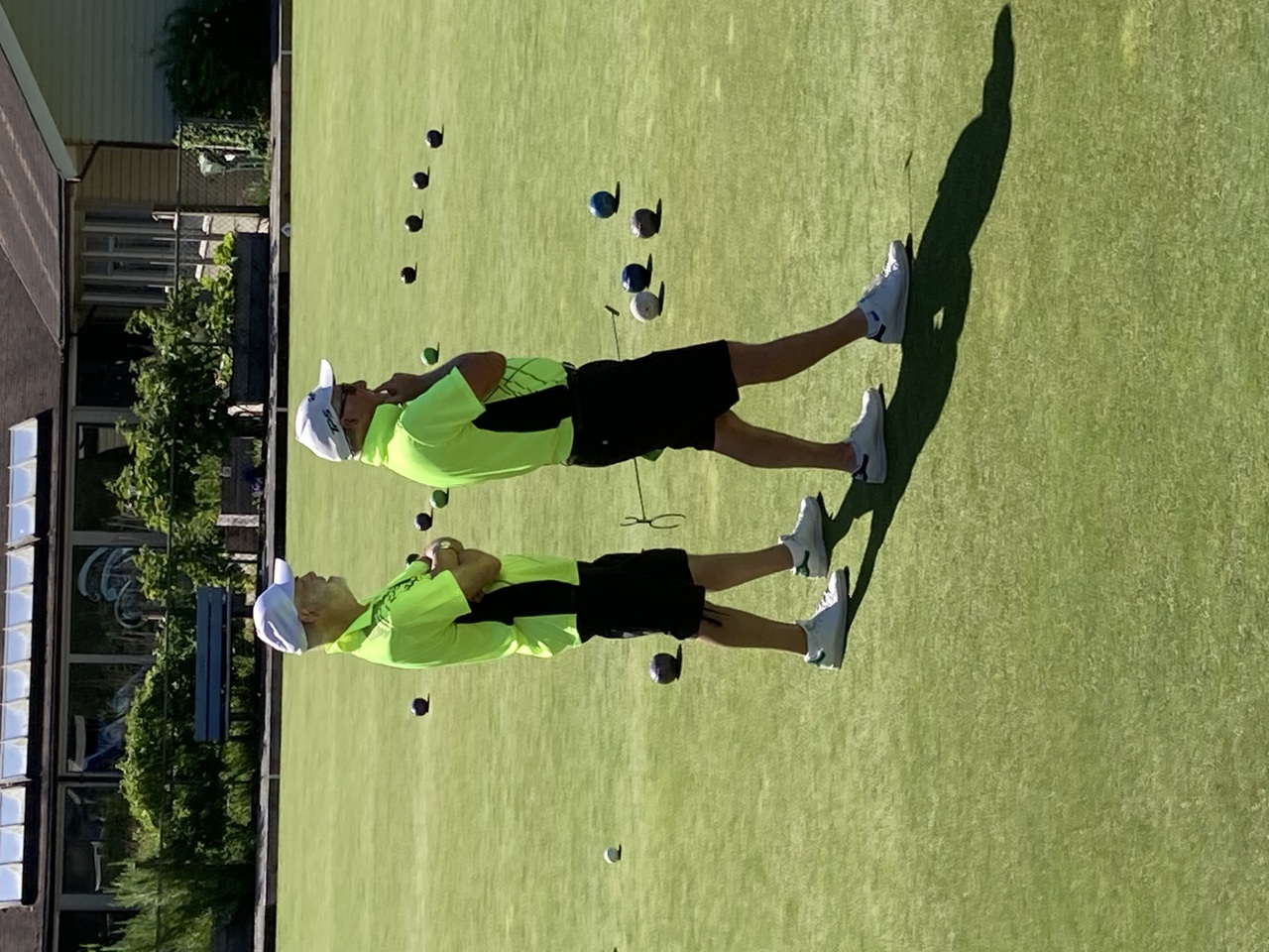 Two men in matching neon green shirts stand on a lawn bowling green observing the game, with scattered bowls visible on the grass. Two men in matching neon green shirts stand on a lawn bowling green observing the game, with scattered bowls visible on the grass.