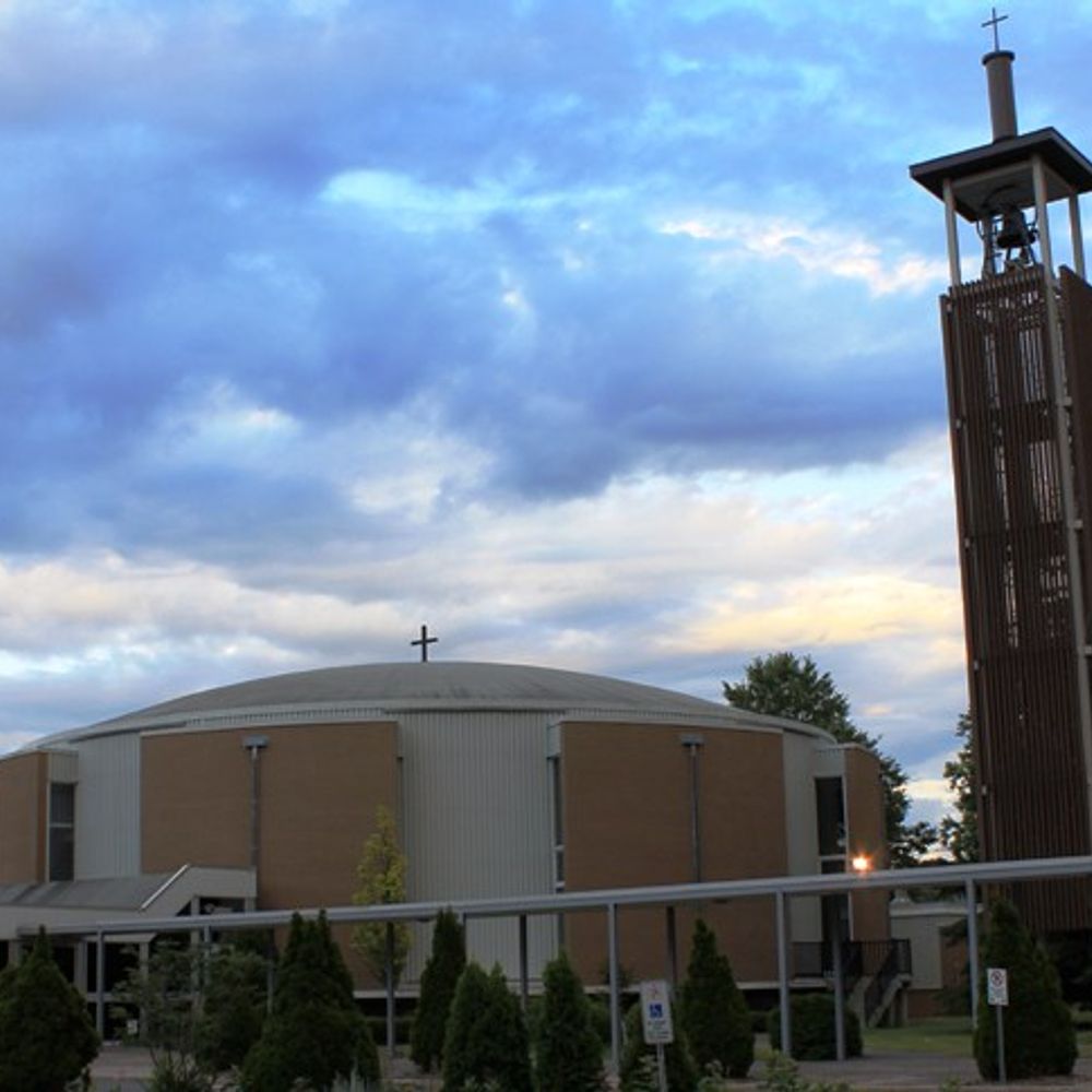 People seated outdoors facing a chapel with a cross in a serene garden setting. People seated outdoors facing a chapel with a cross in a serene garden setting.