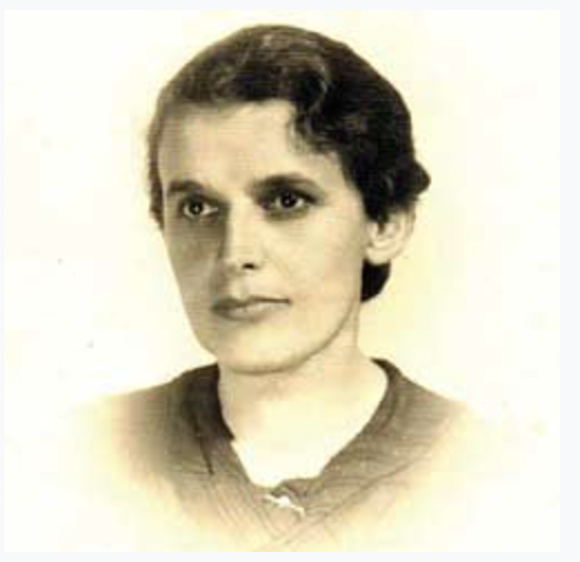 Smiling woman with long brown hair looking out a window, wearing a black top in a softly lit room.
