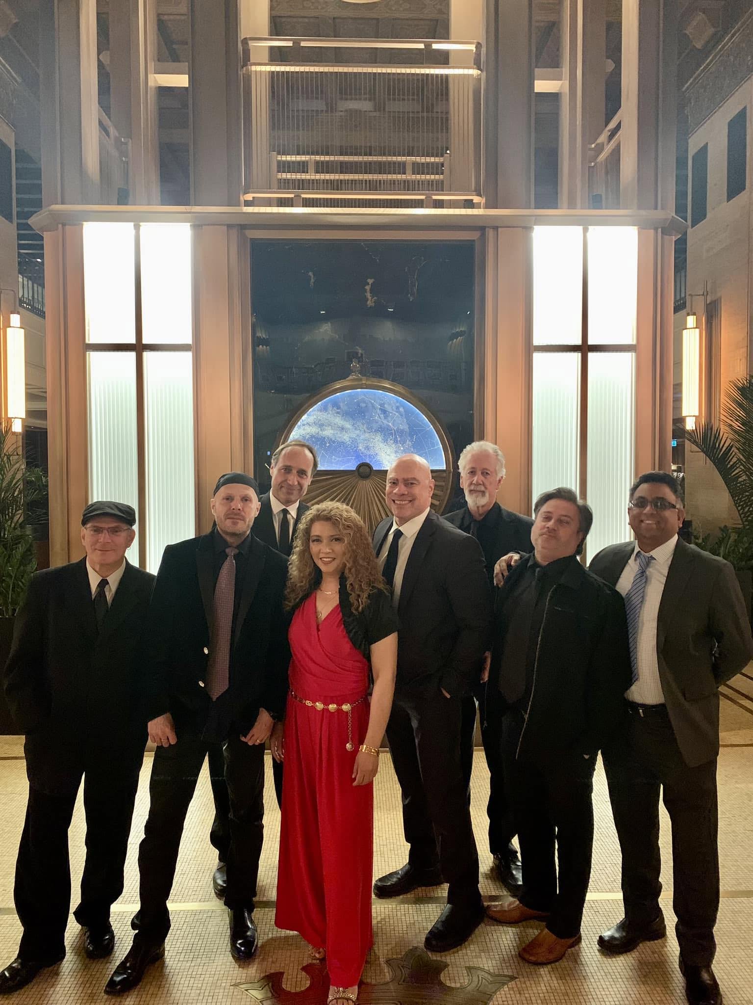 Group of eight formally dressed people posing in an elegant Art Deco lobby with illuminated panels and a circular blue backdrop