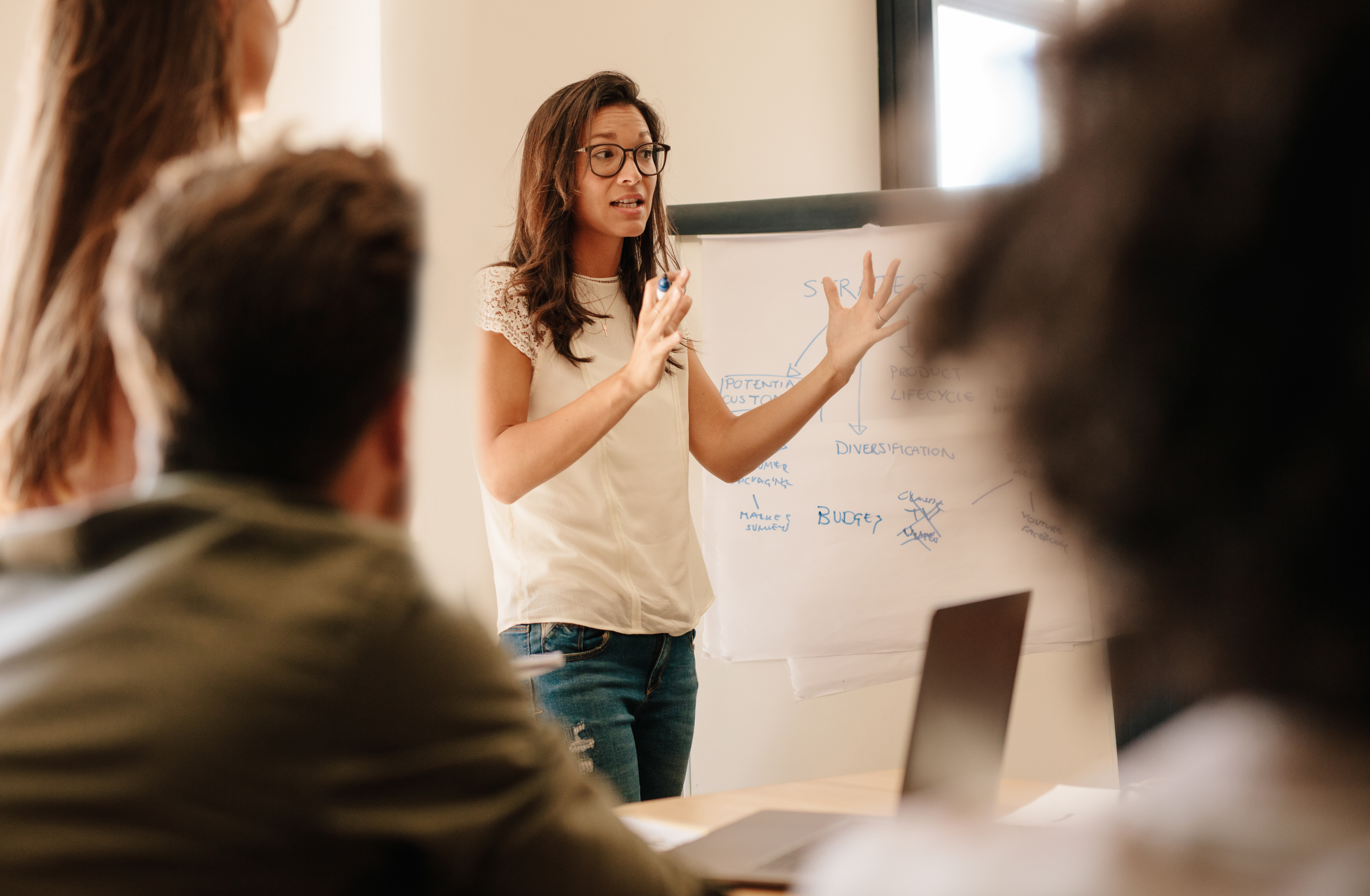 Young woman giving a presentation in an office, pointing to a whiteboard with business strategy diagrams, while team members listen attentively.