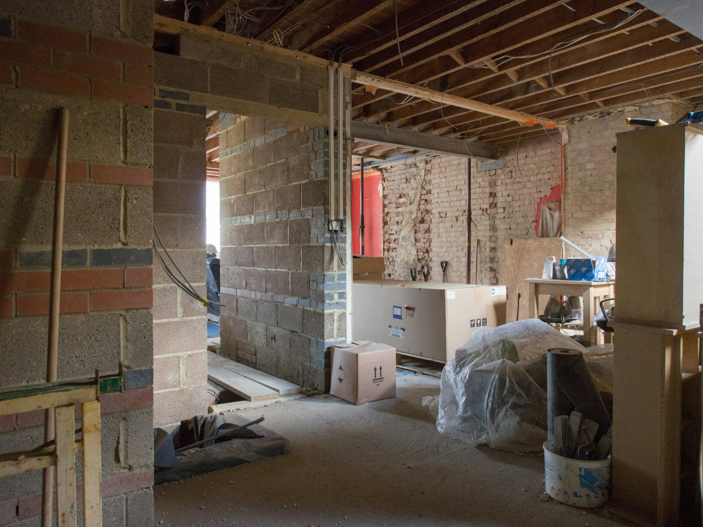 Interior of a construction site in progress showing exposed brick walls and ceiling beams, with construction materials and equipment scattered on the floor.