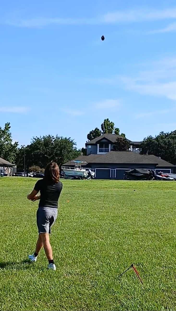Person practicing kick with football on grassy field, houses in background, under blue sky.